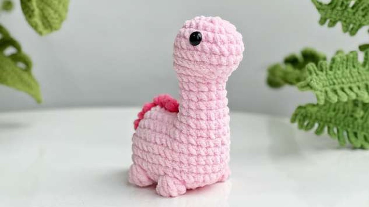 Pink no-sew crochet brachiosaurus with a long neck and dark pink spikes, next to crochet leaves