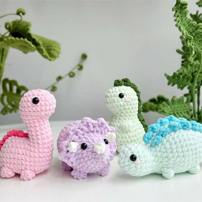 Four finished crochet dinosaurs together: a pink brachiosaurus, a purple triceratops, a pale green T-rex and a mint stegosaurus