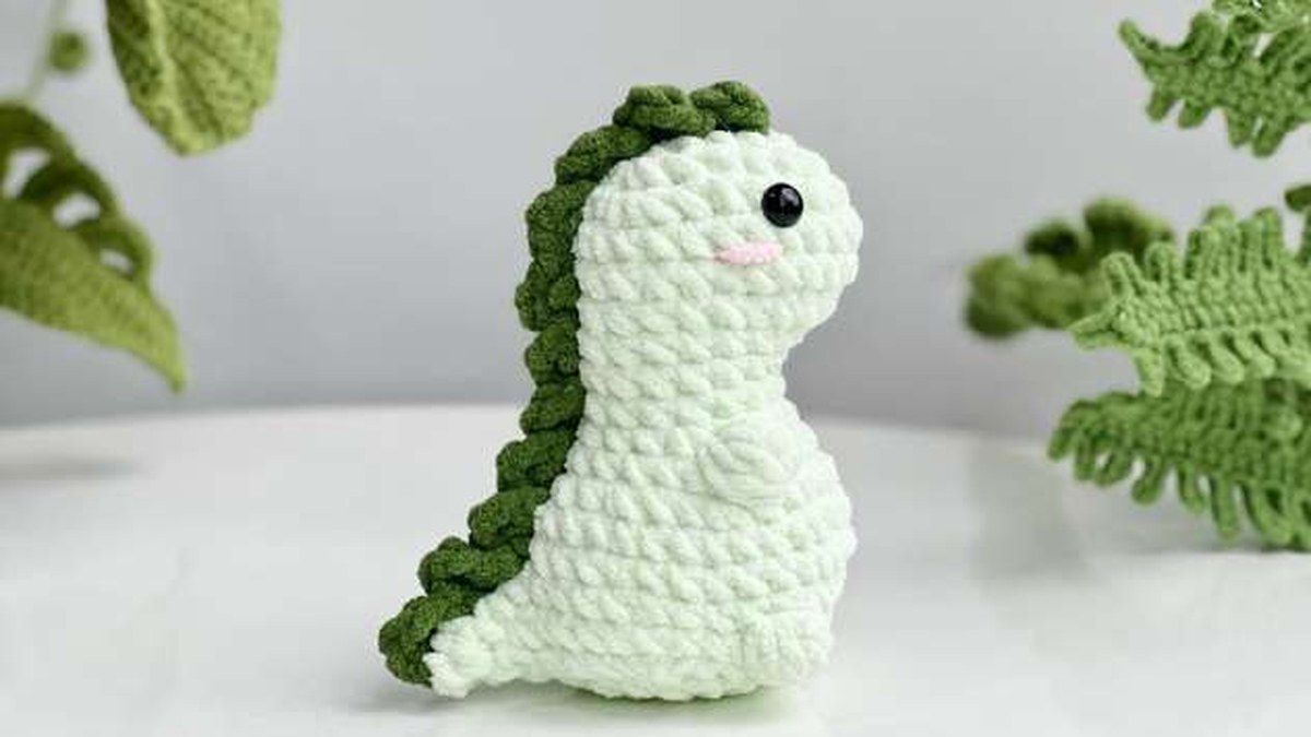 Pale green no-sew crochet T-rex with dark green spikes, next to crochet ferns