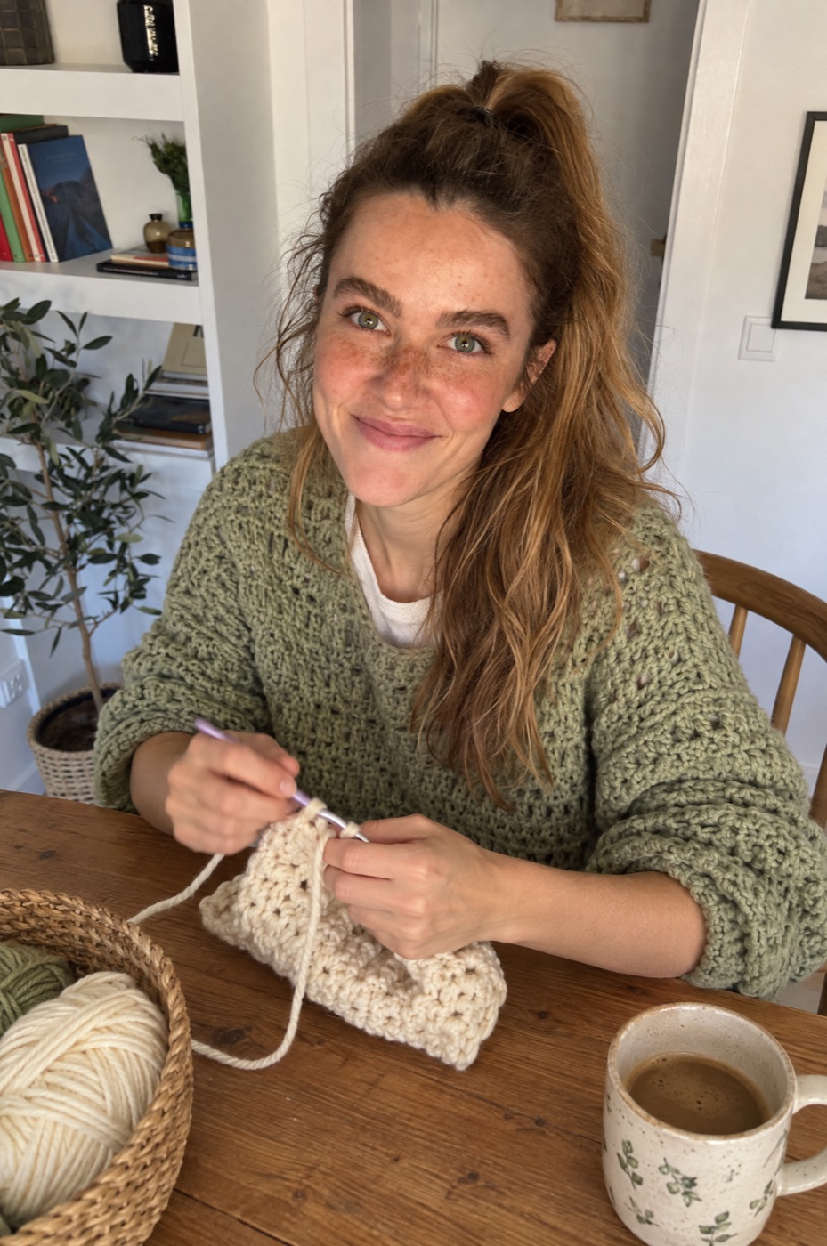 Susie crocheting a cream piece at her kitchen table with a basket of yarn and a cup of coffee